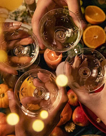 Four hands holding glasses for a toast over a festive table with pumpkins and orange slices, surrounded by soft glowing lights.