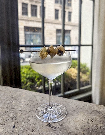 A chilled cocktail in a stemmed glass with a cloudy, pale beverage, garnished with two small cookies on a rod over a windowed bar view.